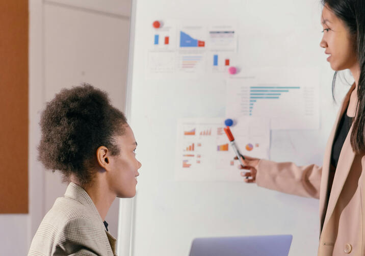 Two women in a meeting, one standing next to a flip chart while the other is seated, discussing governance and compliance.