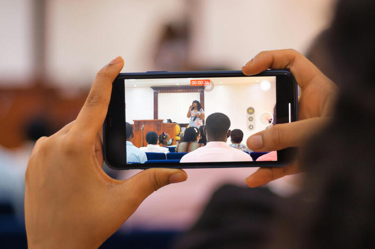 A person recording a video using a smartphone in landscape mode, positioned on a tripod in a naturally lit room. The phone screen shows a live preview of a speaker sharing their story.